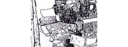 Hier ist eine Zeichnung von Tobias Schulenburg zu sehen. Die Zeichnung ist in schwarz-weiss und man sieht eine Ausschnitt von einem Schreibtisch, auf dem kleine beschriebene Notizbücher, ein Stift, eine Fotokamera liegen und ein Becher mit Stiften steht.
