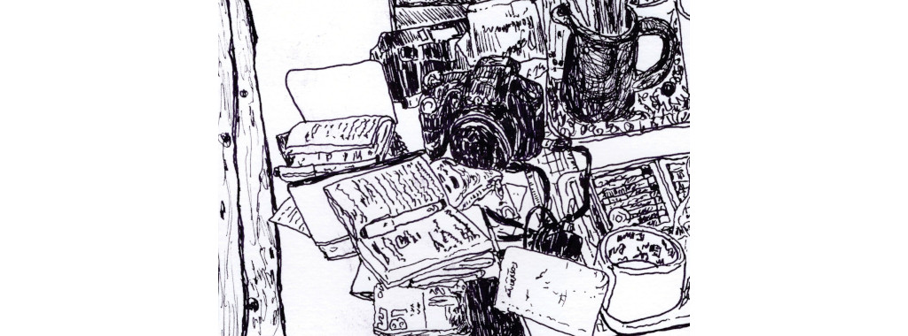 Hier ist eine Zeichnung von Tobias Schulenburg zu sehen. Die Zeichnung ist in schwarz-weiss und man sieht eine Ausschnitt von einem Schreibtisch, auf dem kleine beschriebene Notizbücher, ein Stift, eine Fotokamera liegen und ein Becher mit Stiften steht.